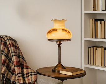 Può includere: Una lampada da tavolo con base in metallo color bronzo scanalato e un paralume in vetro arrotondato con bordo arricciato in un caldo colore crema traslucido. La lampada emette una luce soffusa e presenta un supporto verticale sottile e circolare.