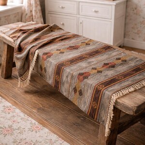 May include: A rectangular woven table runner with a geometric pattern in shades of brown, tan, and rust, fringed at the ends. The runner is draped over a rustic wooden bench, showcasing its texture and design.