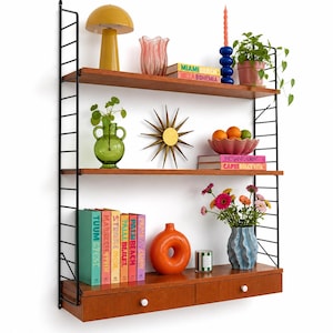 Peut inclure: Une étagère murale de style rétro avec divers objets décoratifs. On y trouve une lampe jaune, un vase rose, des livres, une étoile dorée et un vase de fleurs. Les étagères sont en bois, avec une structure en métal noir. Les livres affichent les textes "MIAMI BEACH" et "IBIZA BOHEMIA".