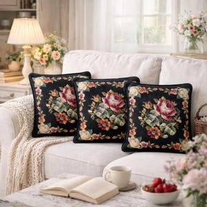 Puede incluir: Tres cojines cuadrados con fondo negro y bordado floral. Cada cojín presenta una gran rosa rosa rodeada de flores coloridas y un borde crema. Los cojines están sobre un sofá blanco.