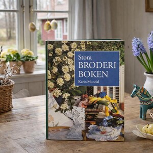 May include: A still-life image featuring a book titled "Stora Broderi Boken" with a floral cover, a basket of pussy willow branches, and a vase of purple hyacinths. Easter eggs and a decorative horse are also present.
