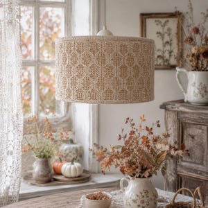 May include: A cylindrical pendant light fixture featuring a beige woven shade with a repeating geometric honeycomb pattern. The shade is suspended by a single cord and has a solid, light-colored circular mounting cap at the top center.