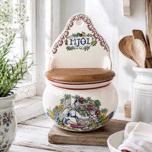 Puede incluir: Un recipiente para harina de cerámica color crema para montar en la pared con tapa de madera. El frente presenta una ilustración colorida de un chef en una cocina y la palabra "MJÖL" en azul. Tiene un borde rojo y detalles florales.