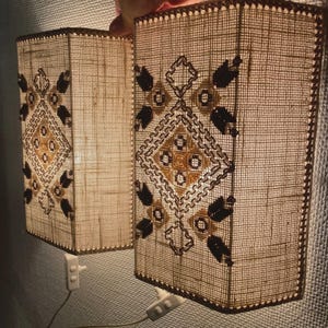 Peut inclure: Deux appliques murales rectangulaires dotées d'un abat-jour en tissu tissé beige clair avec un motif géométrique brodé en brun foncé et jaune doré. Chaque lampe comprend un cordon d'alimentation en plastique blanc avec interrupteur.