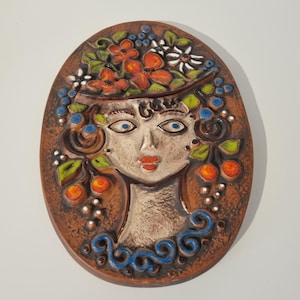May include: An oval-shaped ceramic wall plaque featuring a relief portrait of a person with stylized facial features. The figure is adorned with a crown of orange flowers, green leaves, and blue berries, with additional fruit and scroll motifs.
