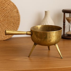 May include: A vintage brass pot with a handle and three legs. The pot has a rounded bowl shape and a golden hue. The handle is attached to the side, and the legs provide stability. The pot is likely used for cooking or decorative purposes.