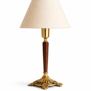 May include: A decorative table lamp with a cream-colored lampshade. The lamp features a brass-colored base and accents, a dark brown wooden stem, and a gold-colored neck. The lamp is designed for indoor use and has an elegant, vintage aesthetic.