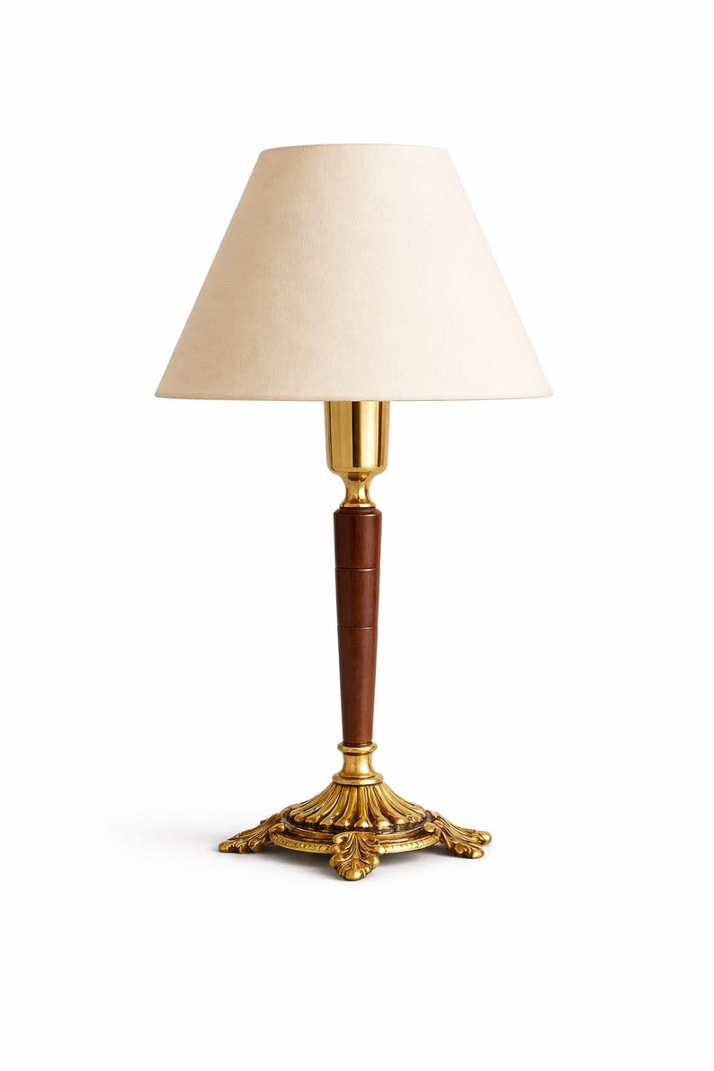 May include: A decorative table lamp with a cream-colored lampshade. The lamp features a brass-colored base and accents, a dark brown wooden stem, and a gold-colored neck. The lamp is designed for indoor use and has an elegant, vintage aesthetic.
