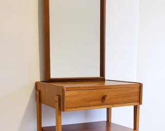 Könnte beinhalten: Ein hölzerner Waschtisch im Mid-Century-Stil mit einem rechteckigen Spiegel. Der Waschtisch hat eine einzelne Schublade mit einem runden Holzknauf, ein unteres Regal und schlanke Beine. Das Holz hat eine warme, natürliche Oberfläche.
