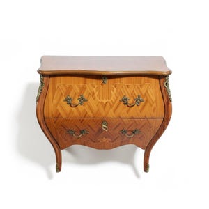 Op de afbeelding: Een houten commode met twee lades, een gebogen voorkant en poten. De commode heeft een lichtbruin bovenblad en een patroon van hout op de lades. Sierlijke messing hardware en decoratieve accenten sieren de commode, wat bijdraagt aan de antieke uitstraling.