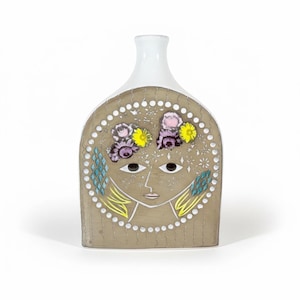 May include: A decorative ceramic vase with a white neck and a tan body. The body features a stylized face with floral accents in pink, yellow, and purple. The design includes geometric and dotted patterns, with yellow and blue accents.