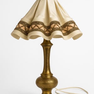 Puede incluir: Una lámpara de mesa de latón vintage con una pantalla de color crema. La pantalla presenta un borde decorativo con detalles en marrón y dorado. La lámpara tiene una base ornamentada y un cable color crema con un enchufe de dos clavijas.
