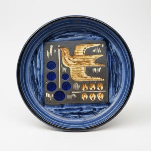 Può includere: Un piatto rotondo in ceramica blu con un bordo strutturato e un design quadrato centrale. Il quadrato presenta un uccello stilizzato in marrone e beige, con cerchi blu e forme a uovo beige. Il piatto ha un bordo blu scuro.