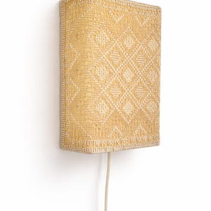 May include: A rectangular wall lamp featuring a textured yellow fabric shade with a repeating white diamond pattern. The lamp has a structured frame and includes a light-colored electrical cord extending from the bottom, coiled at the end.