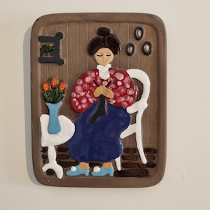 Puede incluir: Una placa de pared de cerámica que representa a una mujer sentada en una silla, tejiendo. Lleva un vestido con estampado rojo y blanco y una falda azul. Hay flores en un jarrón sobre una mesa a su lado.