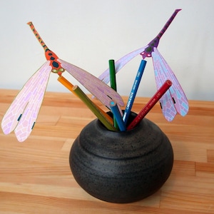 May include: A black ceramic vase with a textured surface holds a collection of colorful pencils and two paper dragonfly decorations. One dragonfly is orange and pink, the other is purple and pink.