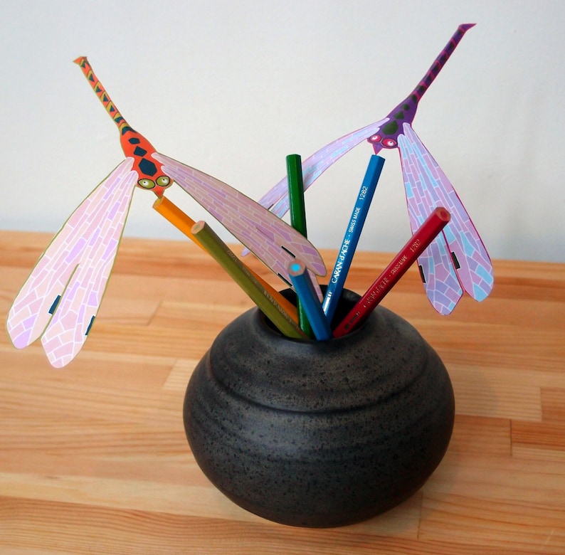 May include: A black ceramic vase with a textured surface holds a collection of colorful pencils and two paper dragonfly decorations. One dragonfly is orange and pink, the other is purple and pink.