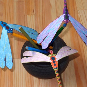 May include: Three colorful dragonfly pencil holders made of paper. The dragonflies are blue, purple, and orange with black eyes. The dragonflies are arranged in a vase with pencils sticking out of them.