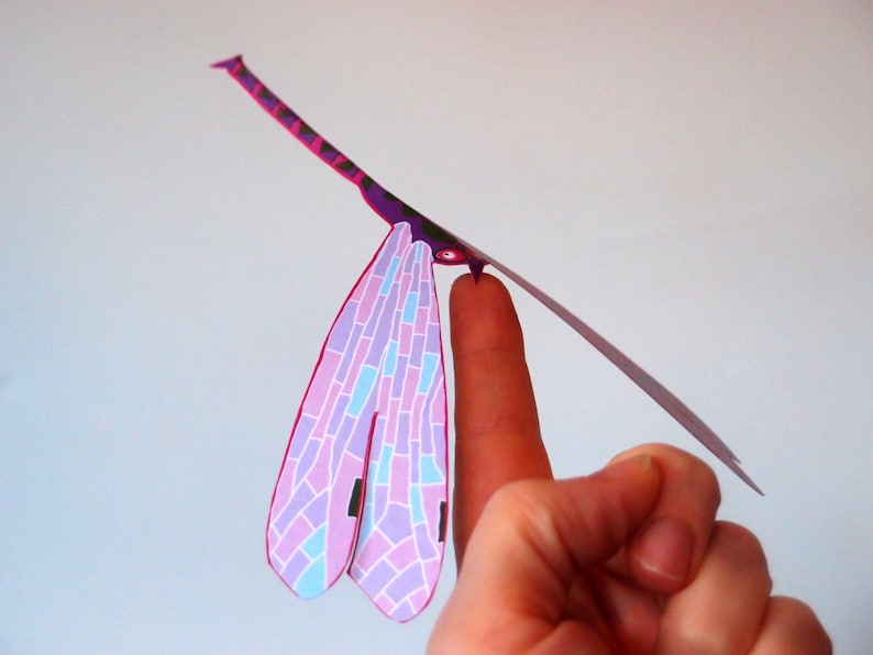 May include: A pink and blue paper dragonfly with a purple body and black accents. The dragonfly is balanced on a finger.