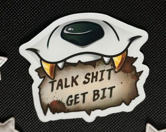 May include: A sticker featuring a stylized monster eye with fangs and the text "TALK SHIT GET BIT." The sticker is white, brown, and gold, with a black background and silver star accents.