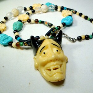 May include: A necklace with a carved wooden mask pendant. The mask has a black and white design and a wide, toothy grin. The necklace is made of various beads, including white, black, brown, and turquoise.