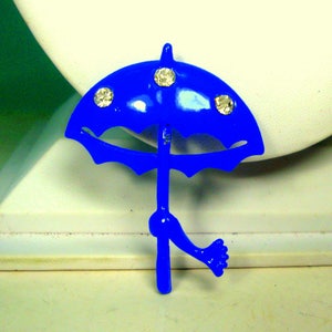 May include: A blue enamel umbrella brooch with three clear rhinestones. The umbrella is tilted to the side and has a leg-like handle.