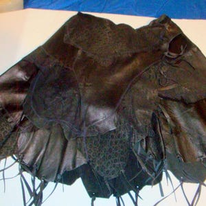 May include: A black skirt constructed from multiple layered panels of leather and textured fabric. The garment features an asymmetrical hemline with dangling leather strips and a combination of smooth and patterned sections throughout the design.