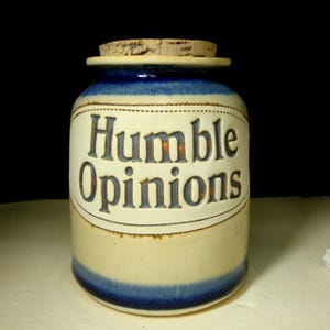 May include: A white ceramic jar with a blue lid and a blue band around the bottom. The jar has the words "Humble Opinions" written on it in a dark gray font.