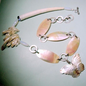 May include: A handcrafted necklace featuring a cream-colored, curved element, connected to a series of iridescent, oval-shaped pieces. A silver butterfly charm and a toggle clasp complete the design. The necklace is made of natural materials.
