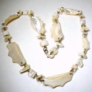 May include: A cream-colored necklace featuring irregularly shaped shell pendants and small square beads. The necklace is strung together with gold-toned metal links, creating a delicate and elegant design. The shell pieces have a natural, iridescent sheen.