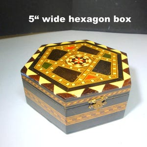 May include: A 5-inch wide hexagon box with a decorative inlaid design. The box features a dark wood exterior with a geometric pattern on the lid, incorporating shades of brown, yellow, red, and green. The box has a brass clasp.