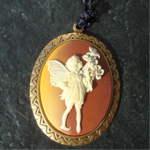 May include: Oval pendant with a cameo of a fairy holding flowers. The fairy is cream-colored, set against a gradient background of orange and brown. The pendant has a gold-colored frame and a dark blue cord.