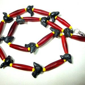 May include: A necklace featuring a repeating pattern of elongated red cylindrical beads, small yellow spacer beads, and dark gray carved animal-shaped beads. The strand is secured with a silver-toned metal lobster claw clasp at the end.