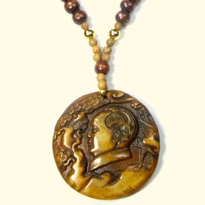 May include: A round, brown, carved wooden pendant featuring a detailed design of a person's face and a tree. The pendant is attached to a brown and gold beaded necklace.