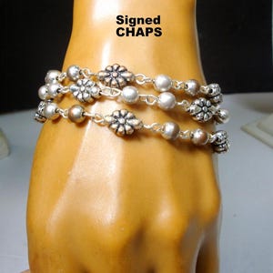 May include: Three-strand silver-tone bracelet with floral and bead accents. The bracelet features a mix of round beads and flower-shaped charms. The text "Signed CHAPS" is visible in the upper left corner.