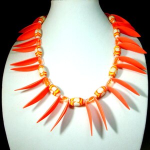 Peut inclure: Un collier orange vif au design unique. Le collier présente des éléments orange allongés entrecoupés de perles ovales blanches, oranges et jaunes. Le collier est présenté sur un mannequin blanc.