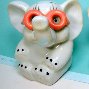 May include: A white ceramic elephant figurine with a long trunk and black dots on its feet. The elephant is wearing large, bright pink, plastic glasses.
