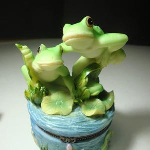 May include: A decorative trinket box featuring two green frogs perched atop lily pads. The box is light blue with a textured design and a decorative clasp. The frogs have brown eyes and are in a playful pose.