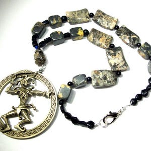 May include: A necklace with a silver-toned jester pendant within a circular frame. The necklace features rectangular and black beads. The jester pendant is silver-toned, and the beads have a mix of colours, including grey, black, and yellow.