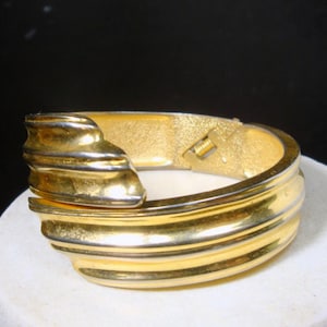 May include: A gold-toned hinged bracelet with a textured, ridged design. The bracelet features a clasp and a decorative, layered element on the front. The metal has a polished finish, reflecting light. The jewelry is set against a neutral background.
