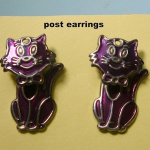 May include: A pair of purple cat-shaped post earrings. The cats are sitting with their tails curled around their bodies. They have white eyes and a white bow tie.