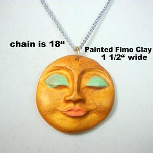May include: A handcrafted pendant necklace featuring a painted Fimo clay sun face with closed green eyes and coral lips. The pendant is 1 1/2" wide and hangs from an 18" silver chain.