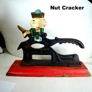 May include: A cast iron nut cracker featuring a painted figure of a person wearing a crown and green clothing, mounted on a rectangular red wooden base. The black metal mechanism has a lever handle and the text Nut Cracker is visible.