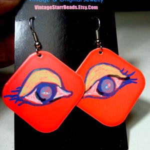 May include: A pair of vibrant, orange diamond-shaped earrings. Each earring features a hand-painted eye design with blue, yellow, and pink details. The earrings have silver-colored fish hook ear wires. The text "VintageStarrBeads.Etsy.Com" is visible.
