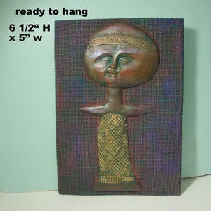 May include: A rectangular wall plaque featuring a relief sculpture of a stylized figure with a large round head and closed eyes. The figure wears a patterned garment and a headband. Text reads: ready to hang, 6 1/2" H x 5" W.