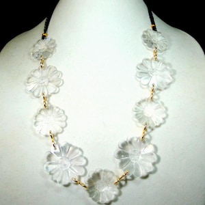 May include: A necklace featuring clear, translucent flower-shaped beads arranged in a series. The floral components are linked together by small gold-toned metal rings, and the piece is suspended from a thin black cord with gold-toned accents.