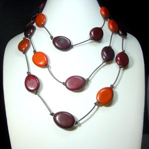 May include: A three-strand necklace with oval beads in shades of orange and burgundy. The beads are strung on a dark cord and displayed on a white mannequin. The necklace is a statement piece, perfect for adding a pop of color to any outfit.