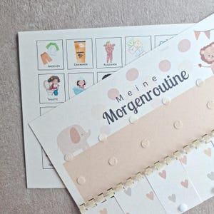 Könnte beinhalten: Eine Morgenroutine-Tabelle für Kinder mit Illustrationen und dem deutschen Text "Meine Morgenroutine". Die Tabelle zeigt Bilder von Anziehen, Auftragen von Sonnencreme und anderen täglichen Aktivitäten. Das Design umfasst ein rosa-weißes Farbschema mit Spiralbindung.