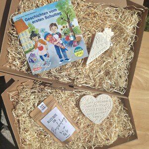Könnte beinhalten: Eine Geschenkbox gefüllt mit Holzwolle, einem Kinderbuch, einer weißen eisförmigen Dekoration mit dem Text "SCHULKIND", einer herzförmigen Dekoration und einem kleinen Beutel mit dem Text "ALLES GUTE ZUR Einschufung". Das Buchcover zeigt Kinder und den Text "Geschichten vom ersten Schultag".