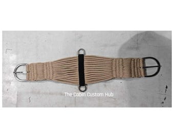 May include: A beige corded horse cinch featuring a central black strap with two metal rings, silver-toned buckles at both ends, and the text The Cabin Custom Hub printed in white below the centre. The design consists of multiple parallel cords.